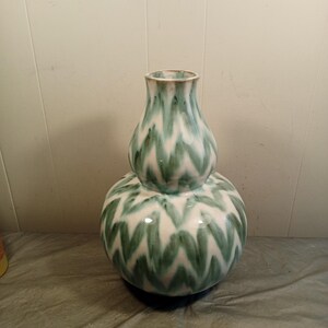May include: A decorative ceramic vase with a unique double-bulb shape. The vase features a white base with a repeating green chevron pattern. The top of the vase has a narrower neck and a green and gold rim. This vase is a stylish home decor item.