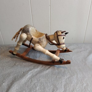 May include: A white wooden rocking horse with a brown leather bridle and a rope tail. The horse is on a wooden rocking base.