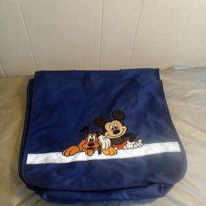 Puede incluir: Bolso mensajero azul marino con una imagen bordada de Mickey Mouse y Pluto. El bolso tiene una franja plateada reflectante en la parte delantera. El bolso está hecho de un material duradero y resistente al agua.
