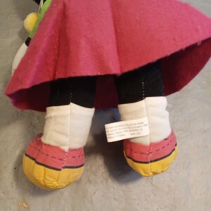 May include: A plush doll featuring a black body, white legs, and red and yellow shoes. The doll has a pink skirt and a tag attached to one leg. The doll's design is reminiscent of a cartoon character.