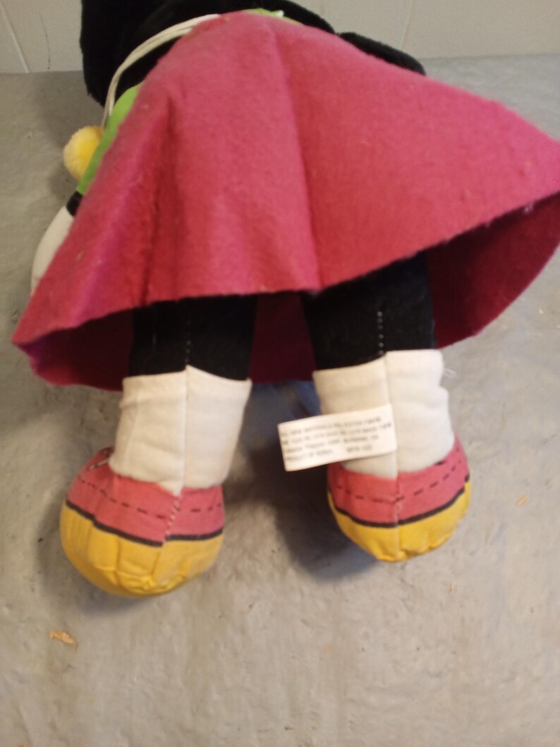 May include: A plush doll featuring a black body, white legs, and red and yellow shoes. The doll has a pink skirt and a tag attached to one leg. The doll's design is reminiscent of a cartoon character.