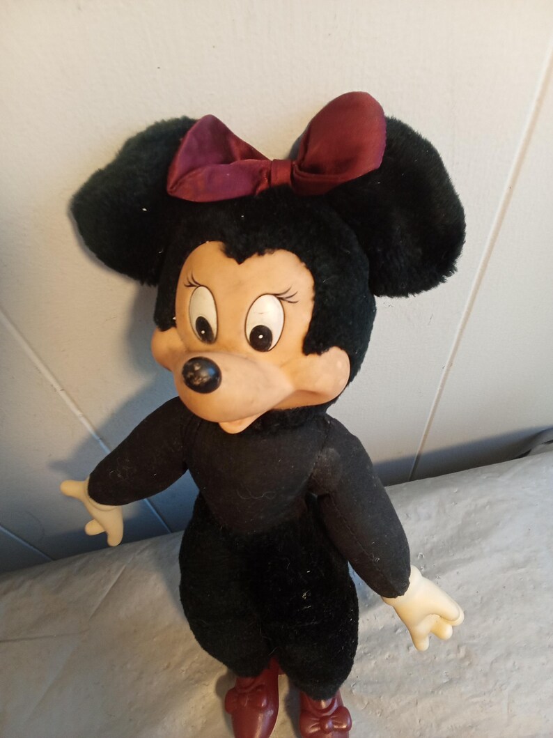 May include: A vintage Minnie Mouse plush doll. The doll has a black body, large black ears, and a burgundy bow. The face is tan with large eyes and a black nose. The doll is wearing burgundy shoes and has white hands.