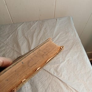 May include: A vintage book with worn, tan-colored pages and a weathered spine. The edges of the pages show signs of age and use. The book's cover is a light brown color, with visible wear and tear.