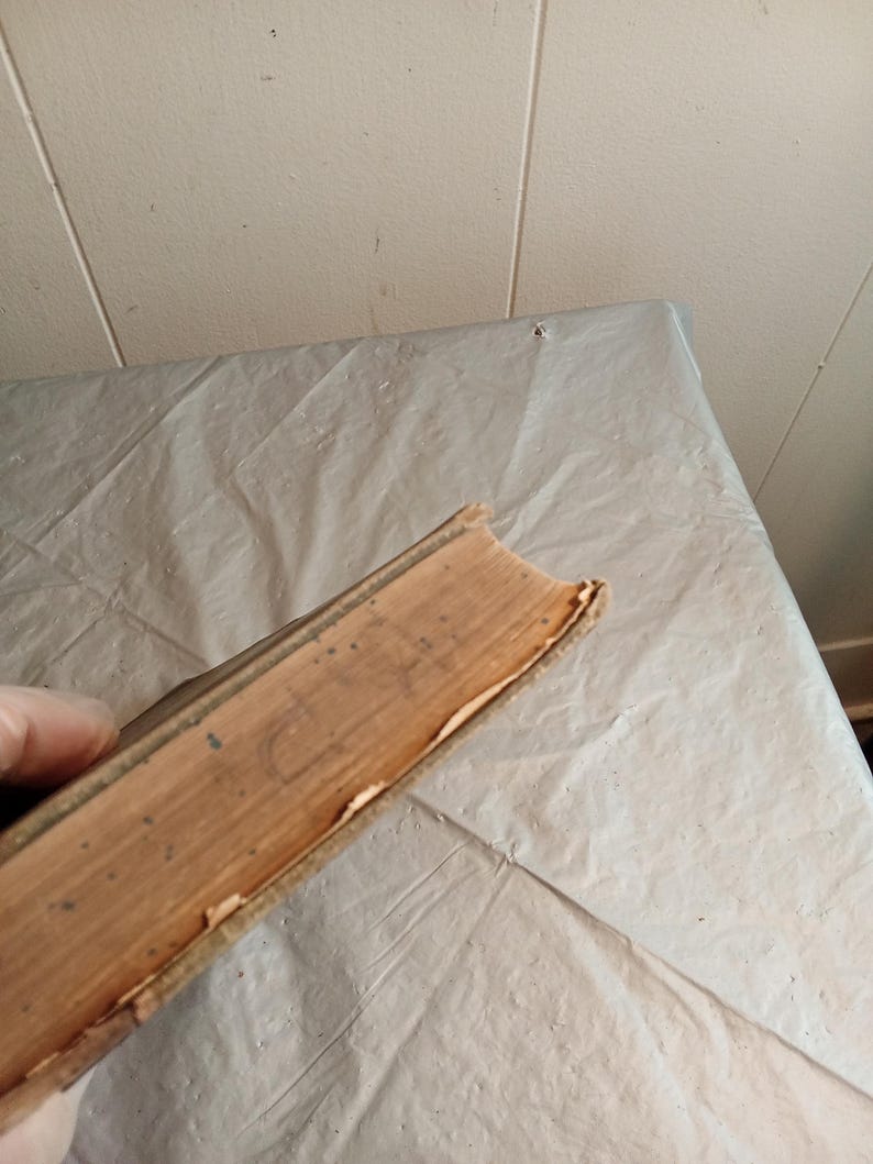 May include: A vintage book with worn, tan-colored pages and a weathered spine. The edges of the pages show signs of age and use. The book's cover is a light brown color, with visible wear and tear.