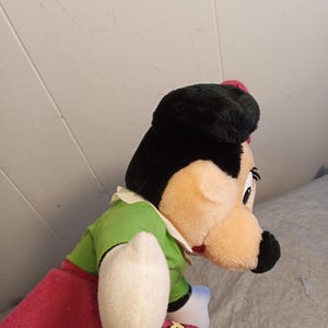 May include: A plush doll of a cartoon character with a black hat, black hair, and a peach-colored face. The doll wears a green shirt, a red skirt, and white gloves. The doll's nose is black, and it has a small red bow.