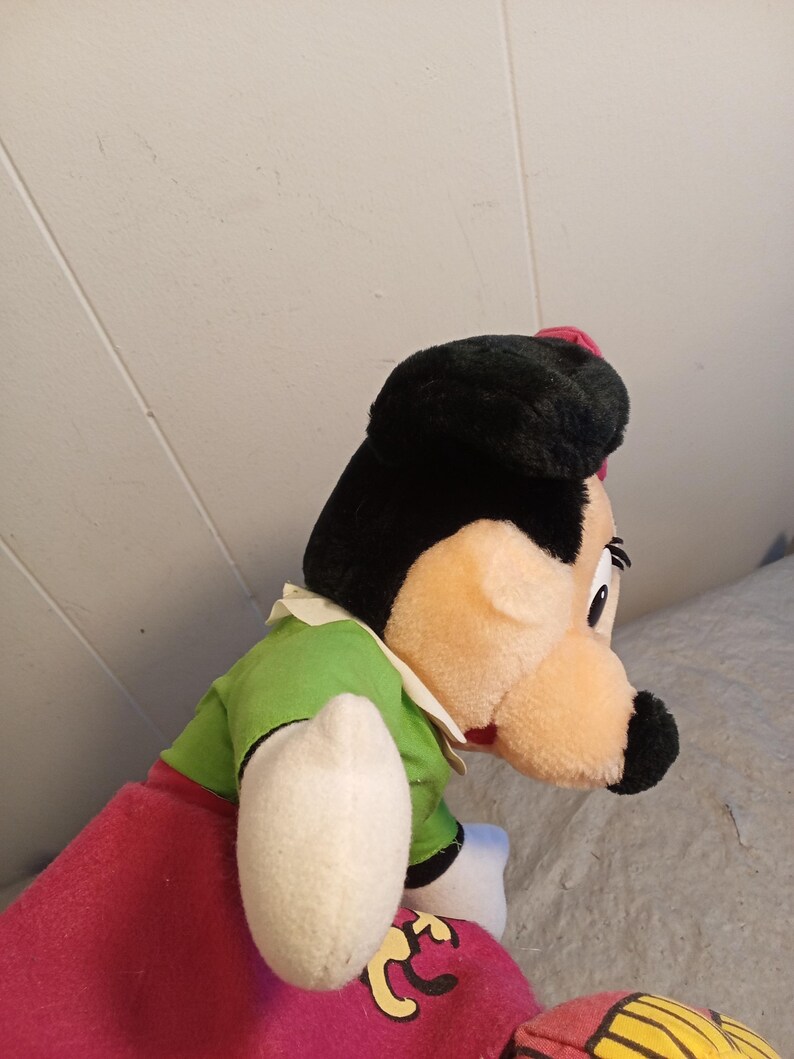 May include: A plush doll of a cartoon character with a black hat, black hair, and a peach-colored face. The doll wears a green shirt, a red skirt, and white gloves. The doll's nose is black, and it has a small red bow.