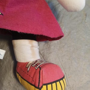 May include: A plush doll's leg and foot are visible, featuring a pink shoe with yellow soles and white laces. The doll wears a white sock and a magenta skirt. The doll's green sleeve and white head are also partially visible.