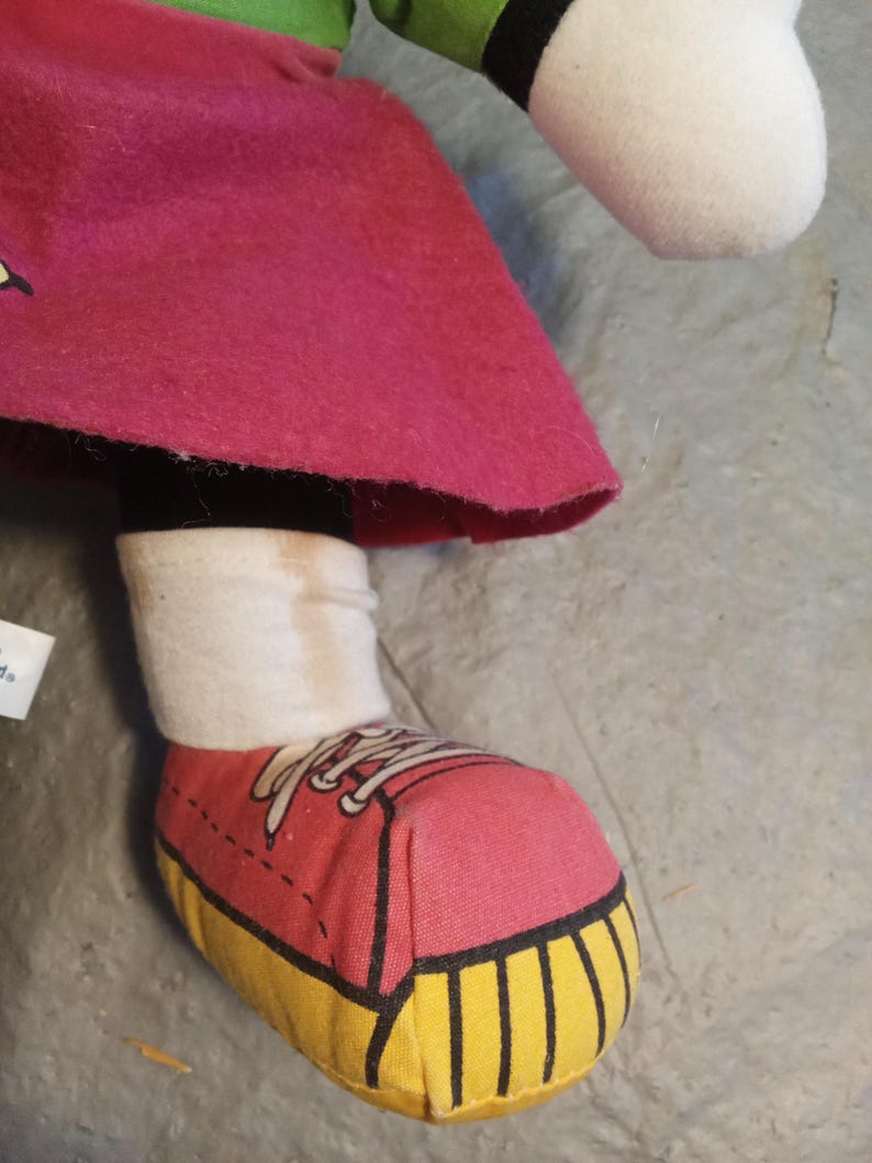 May include: A plush doll's leg and foot are visible, featuring a pink shoe with yellow soles and white laces. The doll wears a white sock and a magenta skirt. The doll's green sleeve and white head are also partially visible.