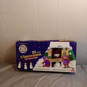 May include: A box set of GE 5 Snowmen specialty lights. The box is dark blue with a winter scene illustration and the text "10 LIGHT". The box has a 3-year guarantee and is for indoor use.