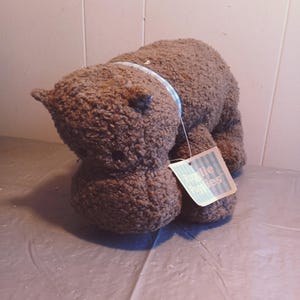 May include: A brown plush bear with a textured, curly fur design. The bear has small ears and a blue and white ribbon around its neck. A tag is attached to the bear.