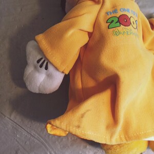 Puede incluir: Un peluche de Mickey Mouse con una chaqueta amarilla con el texto "THE ONE TO 2000 Walt Disney" impreso en la espalda. La chaqueta tiene mangas largas y un cuello. Se ve la mano enguantada blanca de Mickey. El juguete est&aacute; sobre una superficie gris.