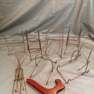 May include: A collection of seven metal display stands in various gold and silver tones, featuring twisted wire designs and folding mechanisms, alongside one solid red painted wooden easel stand for displaying plates, books, or decorative items.