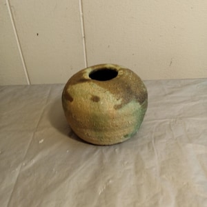Może przedstawiać: Okrągły, ręcznie wykonany ceramiczny wazon z szerokim otworem. Wazon ma nakrapianą kolorystykę brązu, zieleni i beżu, tworząc naturalną, ziemistą estetykę. Sferyczny kształt i unikalne wariacje kolorystyczne sprawiają, że jest to element dekoracyjny.