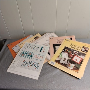 May include: A stack of cross-stitch pattern books with various titles, including "Fall", "From Mother's Hands", "Pattern Pillow", and "Teacher Tributes".