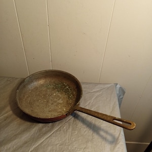 Puede incluir: Una sartén redonda y usada con un mango largo. La sartén tiene un exterior rojo y un interior metálico desgastado. El mango es de color marrón claro con un agujero en el extremo para colgar. La sartén está sobre una superficie gris claro arrugada.