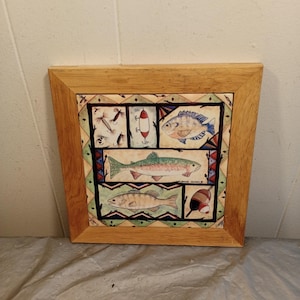 May include: Square wooden framed artwork featuring four fish illustrations and fishing lures. The artwork has a light brown wooden frame. The fish are in different colors and styles. The background has a decorative pattern.