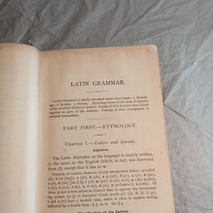 May include: Open book page with the title "LATIN GRAMMAR" at the top. The text discusses etymology, syntax, and prosody, with a section on letters and sounds. The page is aged with a cream-colored background.