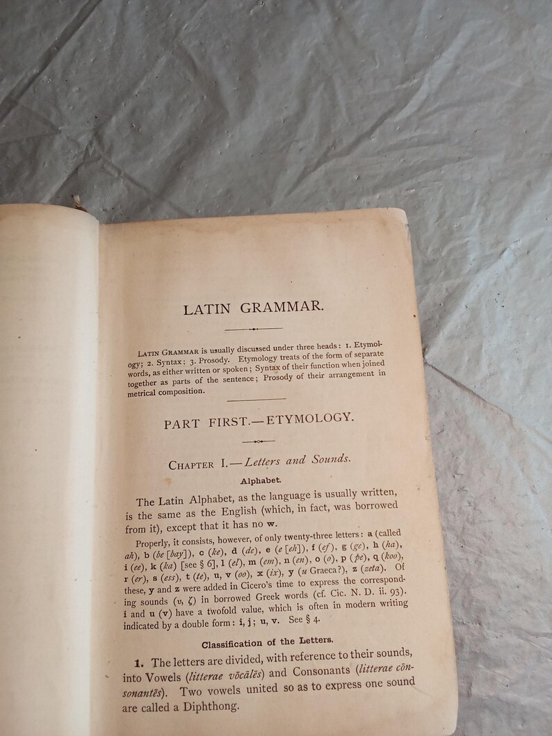 May include: Open book page with the title "LATIN GRAMMAR" at the top. The text discusses etymology, syntax, and prosody, with a section on letters and sounds. The page is aged with a cream-colored background.
