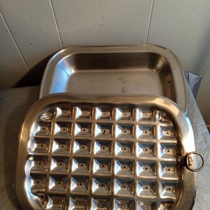 Könnte beinhalten: Eine Edelstahl-Bratpfanne mit Deckel. Der Deckel hat ein Gittermuster aus quadratischen Vertiefungen. Die Pfanne hat eine rechteckige Form mit abgerundeten Ecken und einem Griff. Zum Braten und Backen geeignet.