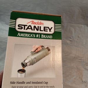 May include: A rectangular cardboard box for an Aladdin Stanley vacuum bottle. The box features a green and beige design with the text Aladdin Stanley America's #1 Brand. It shows a green thermos pouring liquid into a cup with a side handle.