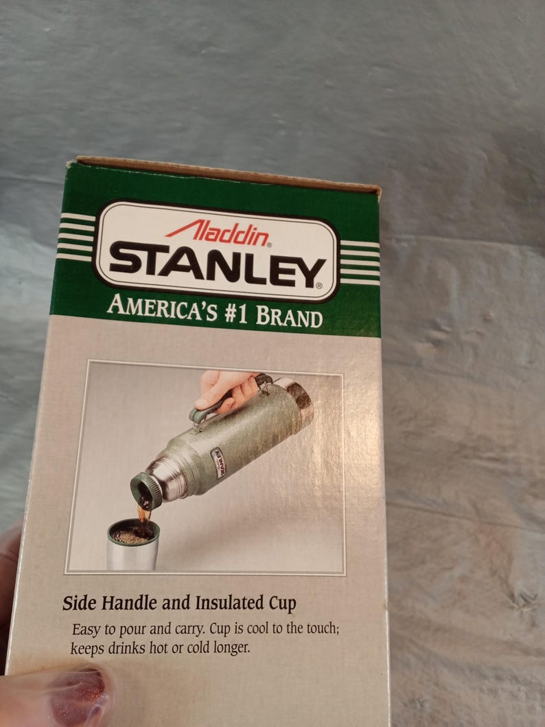 May include: A rectangular cardboard box for an Aladdin Stanley vacuum bottle. The box features a green and beige design with the text Aladdin Stanley America's #1 Brand. It shows a green thermos pouring liquid into a cup with a side handle.