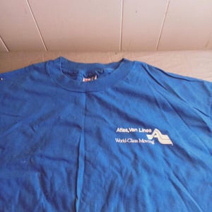 May include: A royal blue t-shirt with a white logo that reads "Atlas Van Lines World-Class Moving" with a stylized mountain range graphic.