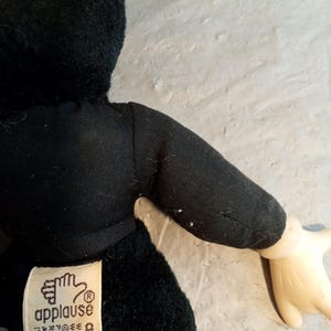 May include: A black plush toy with a black outfit and a tag that reads "applause". The toy has a light-colored hand. The toy is made of soft material.