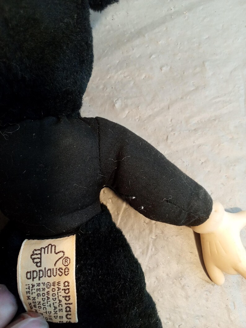 May include: A black plush toy with a black outfit and a tag that reads "applause". The toy has a light-colored hand. The toy is made of soft material.