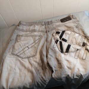 May include: Light gray denim jeans with decorative stitching on the back pockets. The right back pocket features a black and white design. The jeans have a leather patch at the waistband.