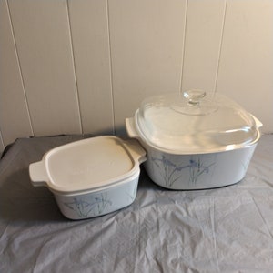 Puede incluir: Dos fuentes blancas para hornear con diseños florales azules. Una fuente tiene una tapa de vidrio transparente y la otra una tapa blanca. Las fuentes están sobre una superficie gris. Probablemente sean para hornear y servir.