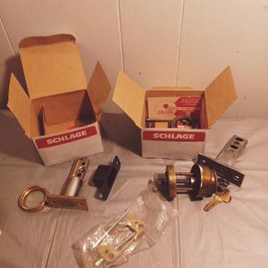 May include: Two open boxes with the Schlage logo, containing a brass door lock set with two keys, a latch, a strike plate, and screws. The lock set is in a clear plastic bag.