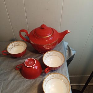 Może przedstawiać: Czerwony ceramiczny zestaw do herbaty z czajnikiem i trzema filiżankami. Czajnik ma pokrywkę i zakrzywiony uchwyt. Filiżanki mają uchwyt i złoty i biały dekoracyjny wzór na obrzeżu. Zestaw stoi na szarej powierzchni.