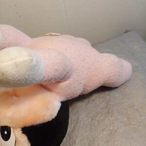 May include: A plush toy featuring a character with a pink head, ears, and hands, and a black face and hair. The toy has white accents on the ears and a large, expressive eye. The toy is resting on a gray surface.