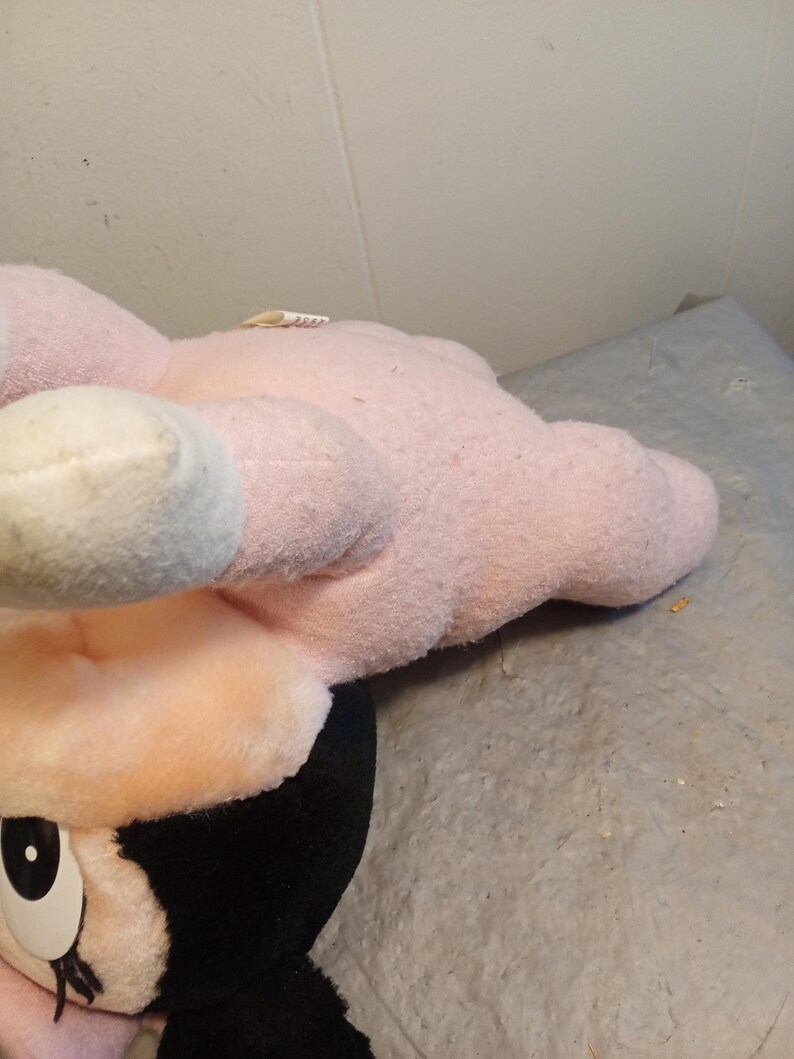 May include: A plush toy featuring a character with a pink head, ears, and hands, and a black face and hair. The toy has white accents on the ears and a large, expressive eye. The toy is resting on a gray surface.