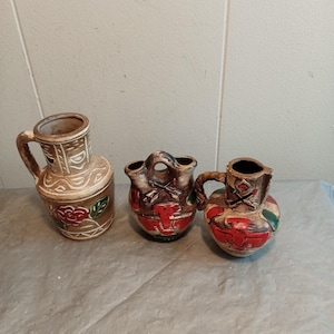 Peut inclure: Trois vases en céramique avec des anses. Les vases sont peints avec des motifs rouges, bruns et verts. Le plus grand vase a un motif de rose. Les deux autres vases ont des motifs géométriques. Les vases sont sur une surface grise.