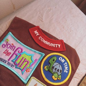 May include: A collection of embroidered patches in various shapes and colors. The patches include text such as "Join the Fun!" and "My Community." A turtle patch reads "On Time." The patches are on a brown fabric.