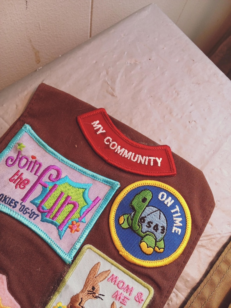 May include: A collection of embroidered patches in various shapes and colors. The patches include text such as "Join the Fun!" and "My Community." A turtle patch reads "On Time." The patches are on a brown fabric.