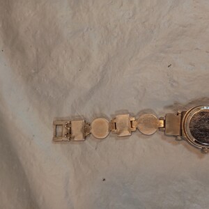 May include: A gold-tone wristwatch featuring a circular case and a link bracelet composed of alternating circular and rectangular metal segments. The back of the watch case is visible, showing a smooth, metallic surface with light surface scratches.