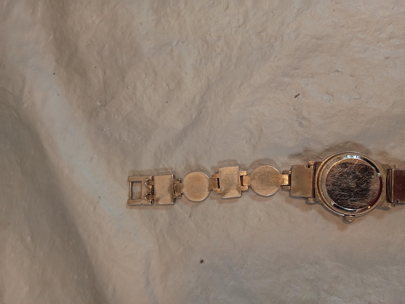 May include: A gold-tone wristwatch featuring a circular case and a link bracelet composed of alternating circular and rectangular metal segments. The back of the watch case is visible, showing a smooth, metallic surface with light surface scratches.