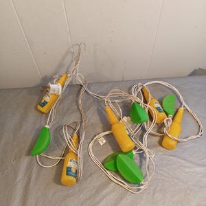 May include: String of decorative lights featuring yellow beer bottle and green lime-shaped ornaments. The lights have a white cord and a yellow plug. The beer bottles have blue and white labels.