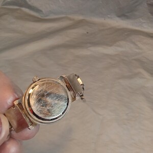 May include: A silver-toned wristwatch featuring a round case with a metallic back plate showing visible surface scratches. The watch includes a matching silver-toned metal link bracelet attached to the case, with a small winding crown on the side.