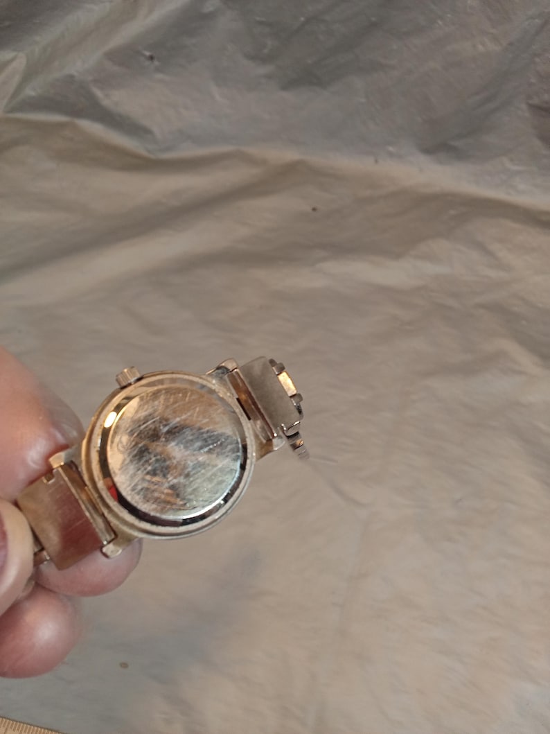 May include: A silver-toned wristwatch featuring a round case with a metallic back plate showing visible surface scratches. The watch includes a matching silver-toned metal link bracelet attached to the case, with a small winding crown on the side.