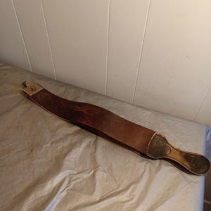 May include: A brown leather strop with a metal end piece and a handle. The strop is curved and appears to be used for sharpening straight razors. The handle has a decorative design.