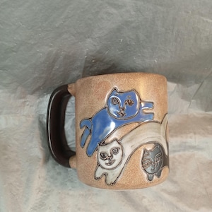 Könnte beinhalten: Eine beige Keramiktasse mit einem dunkelbraunen Henkel. Die Tasse zeigt geprägte Bilder von drei Katzen in Blau, Weiß und Schwarz. Die Katzen sind in einer spielerischen Pose. Die Tasse ist ein einzigartiges Küchenutensil.