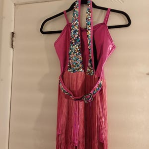May include: A hot pink dance costume with a halter top and spaghetti straps. The bodice is embellished with colorful sequins. A matching belt with a square buckle and fringe skirt complete the outfit. The costume hangs on a black hanger.