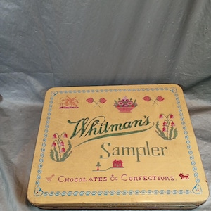 Könnte beinhalten: Eine Vintage-Whitman's-Sampler-Bonbondose. Die gelbe Dose hat ein Kreuzstichdesign mit Blumen- und Flaggenmotiven. Die Worte "Whitman's Sampler Chocolates & Confections" sind in Grün und Rot dargestellt.