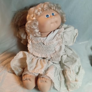 Op de afbeelding: Een stoffen pop met krullend blond wollen haar en helderblauwe ogen. De pop draagt een lichtgroene jurk met een klein wit stippenpatroon, een gesmokt lijfje, mouwen met ruches en een witte kanten rand langs de zoom en de kraag.