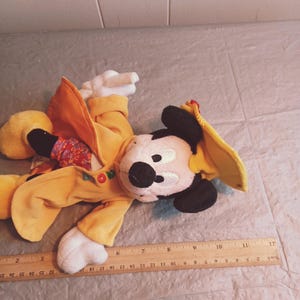 Puede incluir: Un peluche de Mickey Mouse con un atuendo amarillo y un sombrero a juego. El peluche tiene una nariz negra, orejas negras y guantes blancos. Una regla de madera con medidas en cent&iacute;metros est&aacute; en primer plano.