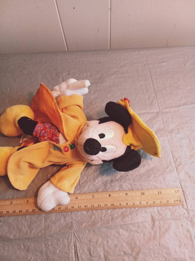 Puede incluir: Un peluche de Mickey Mouse con un atuendo amarillo y un sombrero a juego. El peluche tiene una nariz negra, orejas negras y guantes blancos. Una regla de madera con medidas en cent&iacute;metros est&aacute; en primer plano.