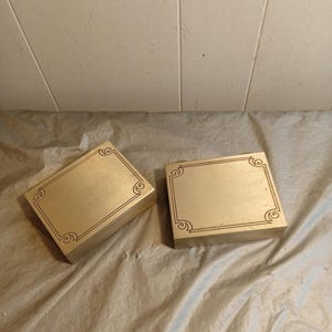 Puede incluir: Dos cajas rectangulares doradas con diseños decorativos en las esquinas. Las cajas están colocadas sobre una superficie arrugada de color claro. Diseño simple y elegante, adecuado para almacenamiento o exhibición.